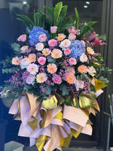 Reflower HK modern luxury grand opening flower stand featuring blue hydrangeas, pink roses, and gold-painted monstera leaves with an elaborate pink and gold base wrapping.