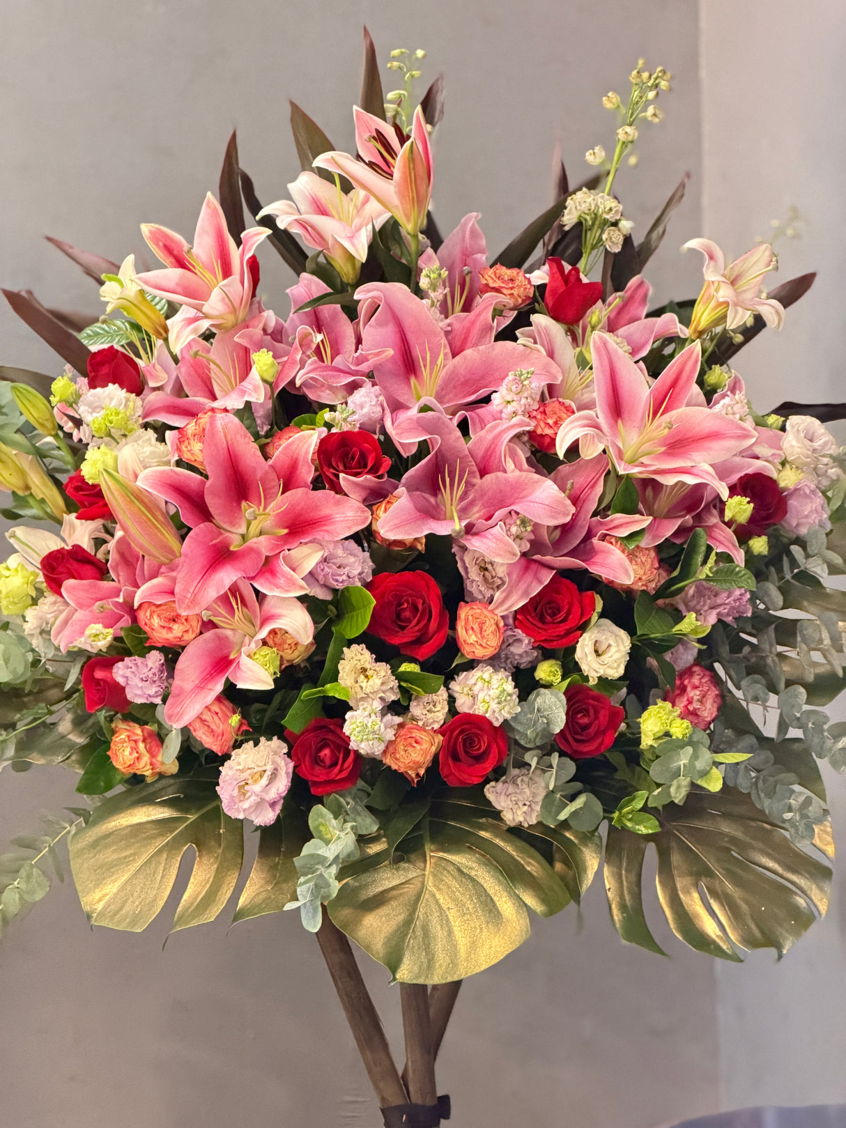 Reflower HK festive luxury grand opening flower stand featuring large pink lilies, red roses, and striking gold-painted monstera leaves at the base, symbolizing golden prosperity.