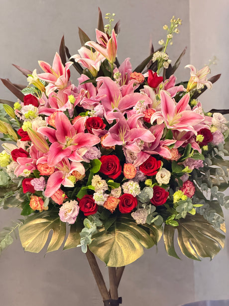 Reflower HK festive luxury grand opening flower stand featuring large pink lilies, red roses, and striking gold-painted monstera leaves at the base, symbolizing golden prosperity.