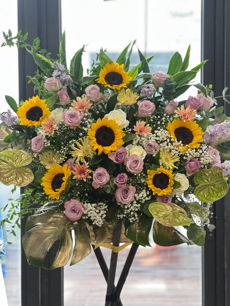 Reflower HK modern grand opening flower stand featuring sunflowers, purple roses, and gerberas, accented with gold-painted monstera leaves and anthuriums at the base.